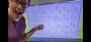 Screenshot image of woman pointing at ASL alphabet chart on tv